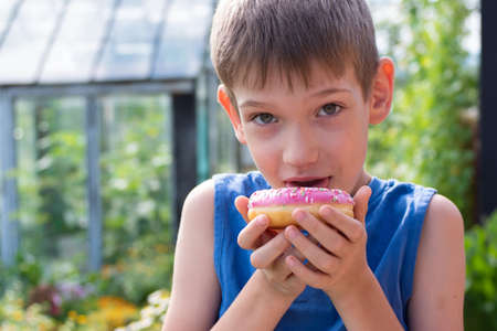 Caucasian boy licks pink glaze from a donut. A child holds a sweet treat in the park. Unhealthy food concept, snacking sweet food. Children love sweet foodの写真素材