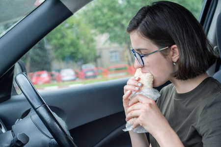 Young beautiful woman eating fast food in the car. Break. Dinner in the car. Front seat. Food concept. Unhealthy food concept, driving, fast foodの写真素材