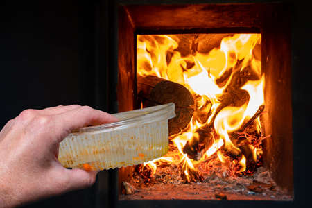 A hand holds a dirty plastic food container next to the fireplace, stove with fire. Recycling, waste, global environmental problems concept. Environmental pollution conceptの写真素材