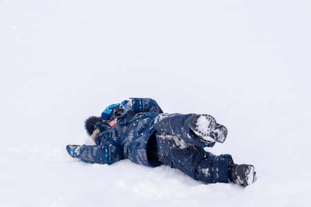 A boy in winter blue clothes lies on the white snow. Outdoor games in the snow. Danger of getting...