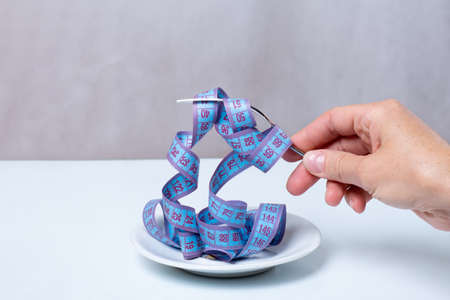 A female hand holds a fork with a coiled measuring tape over an empty plate, close-up, copy space....