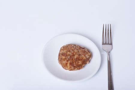 Cooked meat patty on a white plate next to a fork isolated on white background, close-up, copy...