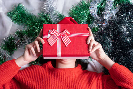 A girl in a red sweater holds a Christmas present in her hand and hides behind the present against a background decorated with green tinsel. Merry Christmas and New Year conceptの写真素材