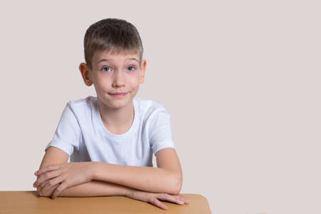 Handsome smart smiling boy sitting at the table and looking forward on a light background, copy space. Home school concept, back to school, distance learningの写真素材