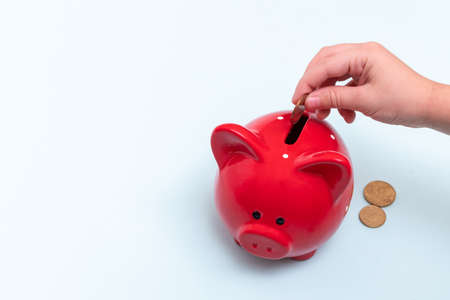 Caucasian female hand puts a coin into a red piggy bank next to lie coins on a blue background, top...