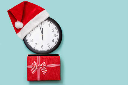 Christmas composition with a classic analog clock in a red Christmas cap and a red gift box with a bow isolated on a blue background, top view, placement from the left, copy space, flat layの写真素材