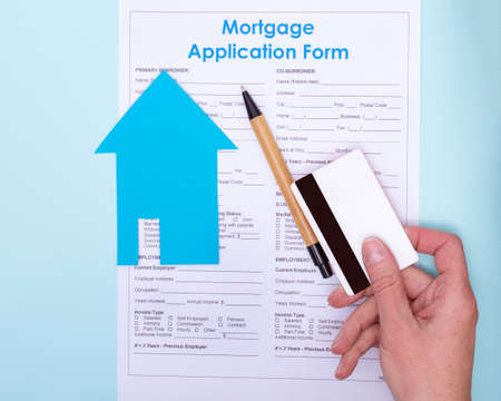 Close-up of a hand giving a bank card to pay for a home mortgage application. Hand with a bank card over a pen and a blue paper house lying on a house mortgage application contractの写真素材