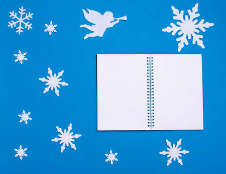 Christmas and new year flat composition with white blank notepad, white angel playing trumpet and christmas snowflakes on blue background, copy spaceの写真素材