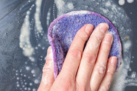 A female hand washes the dishes, a black frying pan with a lilac sponge with foam, close-up, top view. Can be used to illustrate household chores, washing dishes, cleaningの写真素材
