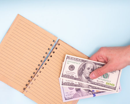 Financial concept. Hand holds dollars over an open blank notepad for writing, close-up, top view, copy space. Can be used in background, layoutの写真素材