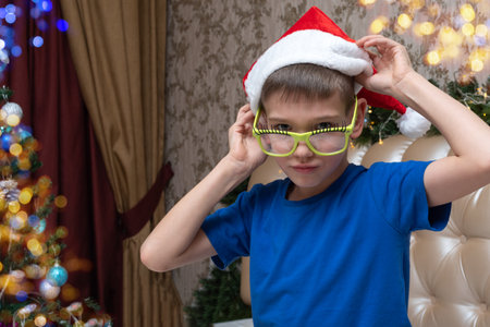 New Year and Christmas traditions. Cute little boy puts on funny glasses and santa hat at home decorated for Christmas. Preparing for a Merry Christmas and a Happy New Yearの写真素材