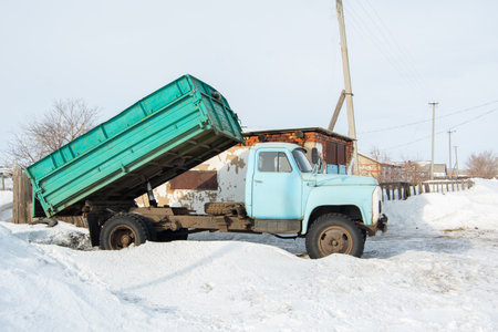 Heavy old blue dump truck with raised body close-up on the background of a snowy road, side view. Mining equipment. Quarry equipment. Unloading truck on a snowy winter roadの写真素材