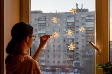 A teenager girl draws snowflakes with white paint on the window, decorates the window for Christmas, New Year at home, rear view. Waiting for winter, miracle, Christmas conceptの写真素材