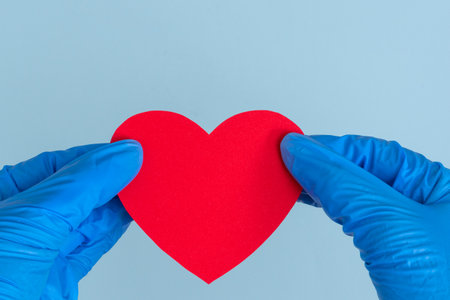 Valentine's day during the coronavirus pandemic. Two hands in blue medical gloves holding a model in the shape of a red heart on a blue background, copy space, top viewの写真素材