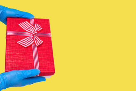 Safe delivery of gifts concept. Hands in blue gloves hold a red gift box with a bow on a trendy...
