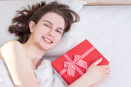 Birthday morning. Lying on the bed happy smiling girl holding a red gift box at home, top view, copy space. Can be used for wedding day, Christmas, Valentine's Dayの写真素材