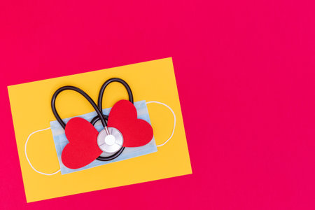 Black heart shaped stethoscope on medical protective mask and red two paper heart shape on trendy yellow Illuminating and red background, top view, copy space. Health care and cardiology conceptの写真素材