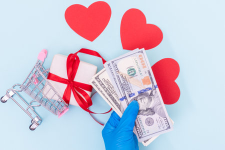 Safe shopping concept. A hand in a blue medical glove holds cash dollars over a shopping cart with a gift box and red love hearts on a blue background, copy space, flat lay, top viewの写真素材