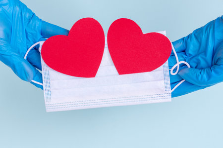 Hands in blue gloves hold a medical mask with two paper love red hearts on a blue background, close up, copy space. Protection against coronavirus on valentine's day concept, symbol of love and safetyの写真素材