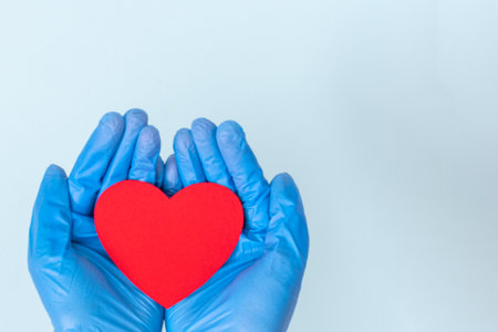 Two hands in medical blue gloves holding a red heart shape on a blue background, copy space. Love,...