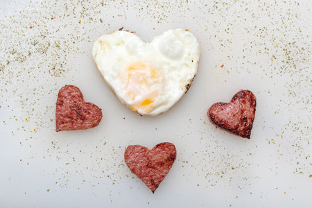 Heart shaped scrambled eggs with heart shaped sausages, herbs and spices on a white plate, top view....