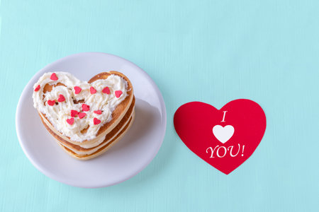 Valentines day creative breakfast. Homemade heart shaped pancake decorated with white cream with red hearts on a white plate on a blue background with a red heart with the inscription I love youの写真素材