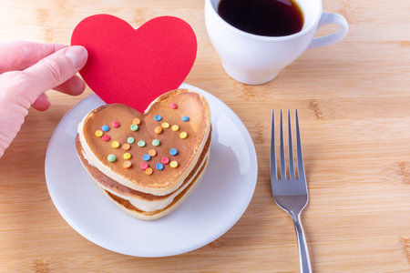 Hand holds a red blank heart shape next to homemade heart shaped pancakes with sugar decoration on a white plate, fork, mug with coffee or cocoa, close-up. Breakfast for Valentines Day Conceptの写真素材