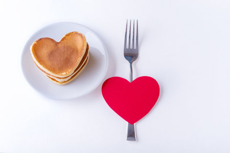 Homemade heart shaped pancakes on a white plate, fork and red blank heart shape on a white background, top view, copy space. Postcard for Valentines Day, Mothers Day, Birthdayの写真素材
