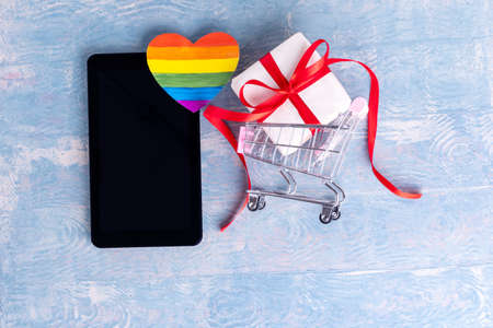 Online shopping for Valentines Day LGBT concept. Online congratulations concept. Gift box in shopping trolley, tablet or smartphone and painted paper LGBT heart shape on blue backgroundの写真素材