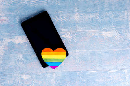 Online congratulations LGBT concept. Blank black screen tablet or smartphone and painted paper LGBT heart shape on blue background, copy space, flat layの写真素材