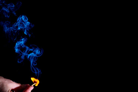 Blue curly smoke from a lit match in a holding hand isolated on a black background. Abstract...