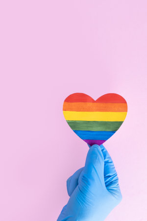 A hand in a protective blue glove holds a rainbow paper heart on a pink background, vertical frame. Safe Valentines Day 2021 concept. LGBTQL conceptの写真素材