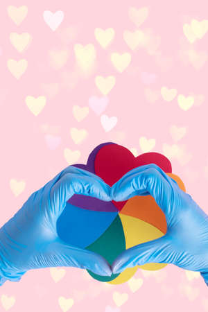 Two hands in protective blue gloves show a heart shape over an iridescent paper circle on a pink...