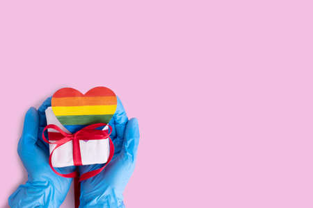Hands in protective blue gloves hold a homemade rainbow paper heart and a gift box with a red ribbon on a pink background, copy space. Safe LGBTQL conceptの写真素材