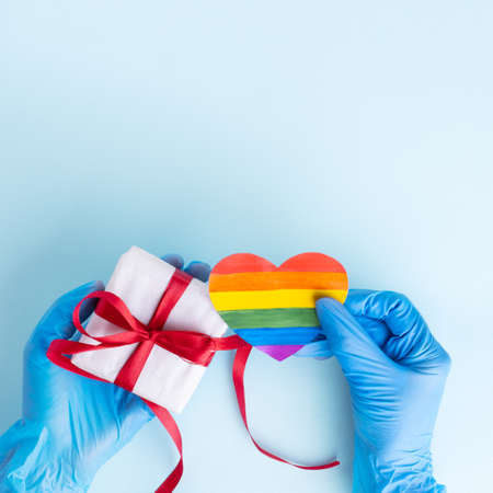 Hands in protective blue gloves hold a homemade rainbow paper heart and a gift box with a red ribbon on a light blue background, copy space. Safe Valentines Day 2021 conceptの写真素材