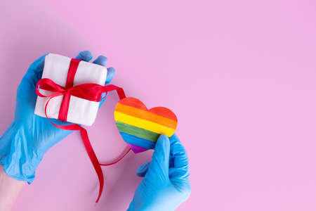 Hands in protective blue gloves hold a homemade rainbow paper heart and a gift box with a red ribbon on a pink background, copy space. Safe Valentines Day 2021 concept. LGBTQL conceptの写真素材