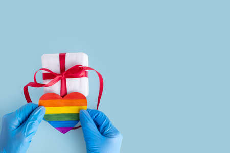 Hands in protective blue gloves hold a homemade rainbow paper heart and a gift box with a red ribbon on a light blue background, copy space. Safe Valentines Day 2021 concept. LGBTQL conceptの写真素材