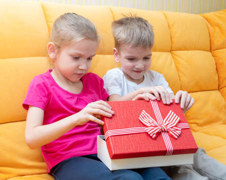 Caucasian attractive little brother and sister sitting on the sofa at home open a red gift box. The boy congratulates the girl on her birthday, Valentines Dayの写真素材