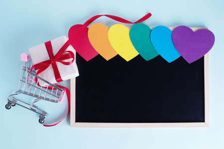 Gift box in shopping carts, multicolored rainbow colored paper hearts and a blank board with copy...