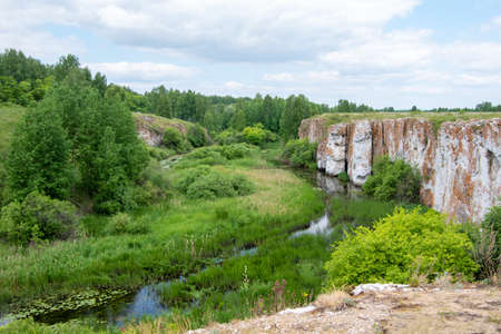 Sunny summer landscape with ravine, swamp, forest and rocky coast. White cumulus clouds in the sky. Bright green marsh grass and small trees by the riverの写真素材
