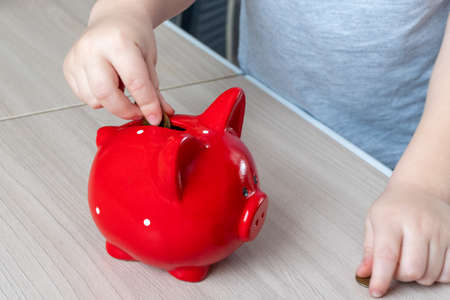 Childrens hand puts a coin in a red piggy bank on a wooden background, top view, copy space. Money saving conceptの写真素材