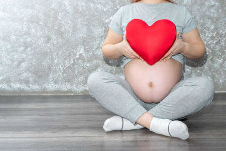 Pregnancy, motherhood, people, love and expectation concept - close-up of a pregnant woman holding a red heart shape pillow at home. A pregnant woman is expecting a baby. Mothers day conceptの写真素材