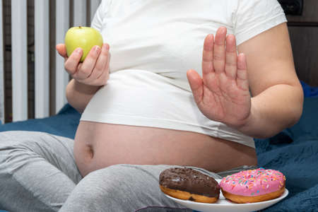 Close-up of a pregnant woman holding a green apple in her hands, refusing donuts. Healthy food...