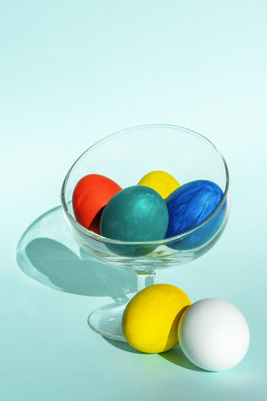Hand-painted colorful Easter eggs in a transparent glass bowl on a light blue background, vertical frame, copy spaceの写真素材