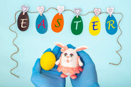 Garland of paper eggs with the inscription EASTER and hands in medical gloves holding a yellow egg...