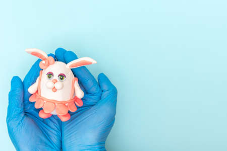 Hands in protective medical gloves holding a homemade Easter bunny in a pink skirt on a blue background.の写真素材