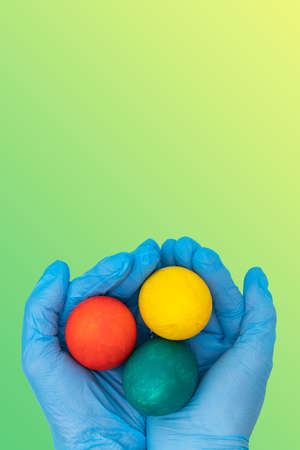 Easter vertical card 2021. Hands in protective medical gloves holding colorful Easter eggs on a gradient spring yellow-green background. Happy safe Easter concept. New norma conceptの写真素材
