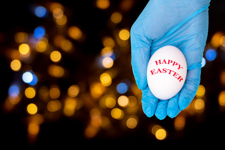 A hand in a protective medical glove holds or gives a white egg with the inscription HAPPY EASTER on a black background with bokeh, copy space. Happy and safe easter conceptの写真素材