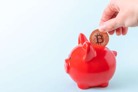 A hand puts a bitcoin coin in a red piggy bank on a blue background, close-up, copy space....