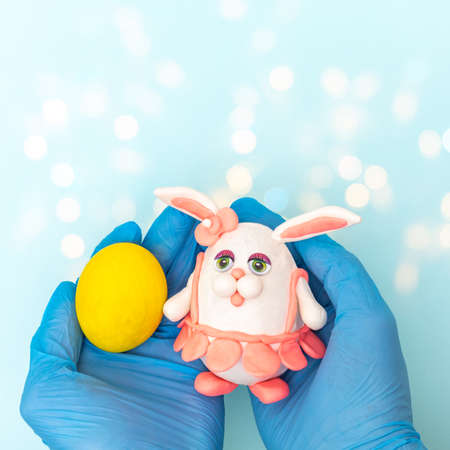 Hands in protective medical gloves holding a painted yellow Easter egg and a homemade Easter bunny...